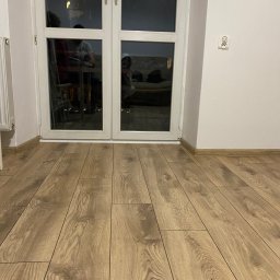 FixPoint - Jasne panele podłogowe ułożone wzdłuż białych drzwi balkonowych. Na ścianie widoczne gniazdko elektryczne. W tle odbicie osób w szybie.