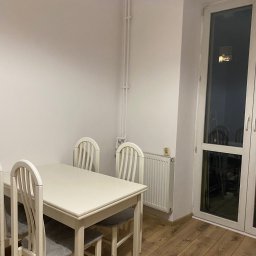 FixPoint - Jasne wnętrze jadalni po remoncie: biały stół z krzesłami, drewniana podłoga, grzejnik i okno balkonowe. Minimalistyczny design, świeżo odnowione ściany.