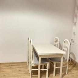 FixPoint - Jasne wnętrze z drewnianą podłogą, białymi ścianami i białym stołem z krzesłami. Widoczny grzejnik i rury przy ścianie. Minimalistyczny wystrój.