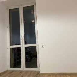 FixPoint - Jasne panele podłogowe w pokoju z białymi ścianami i oknem balkonowym. Widoczne gniazdko elektryczne. Minimalistyczny wystrój.