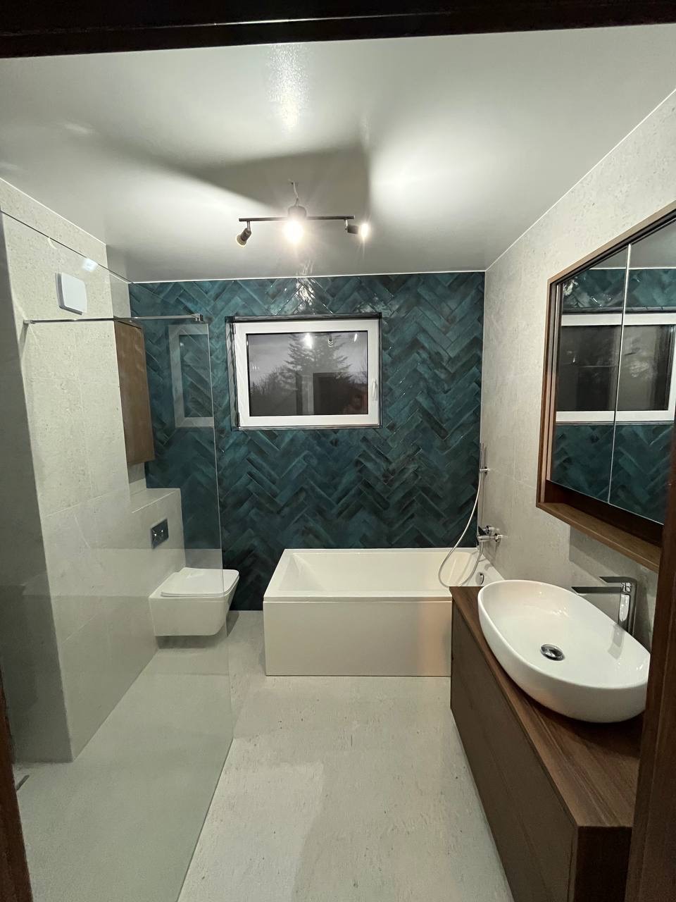 Nowoczesna łazienka z turkusową ścianą ułożoną w jodełkę, białą wanną i umywalką nablatową na drewnianej szafce. Minimalistyczne oświetlenie sufitowe.