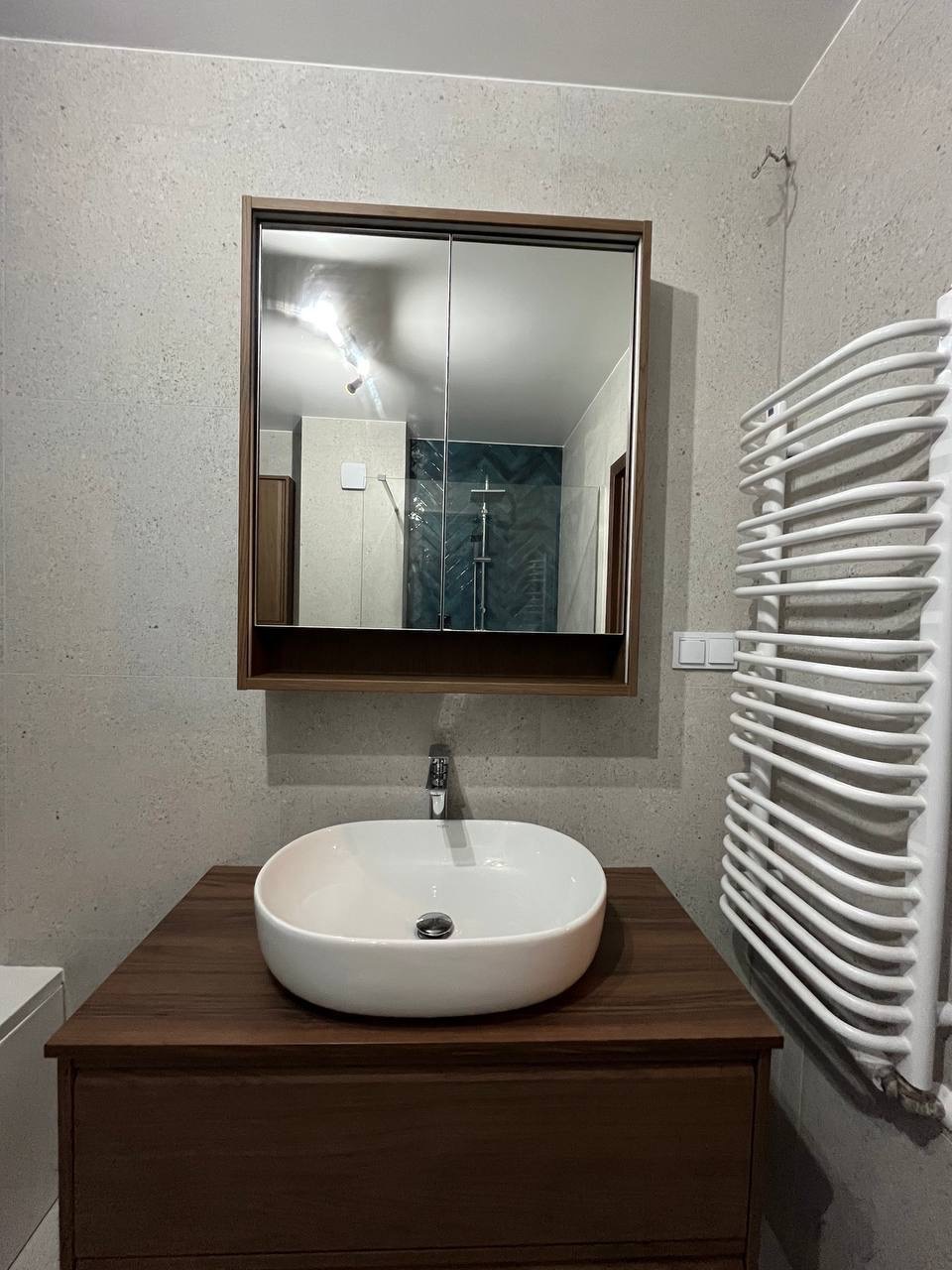 Nowoczesna łazienka z umywalką nablatową, szafką z ciemnego drewna i lustrem w zabudowie. Widoczny grzejnik drabinkowy oraz szare płytki na ścianach.