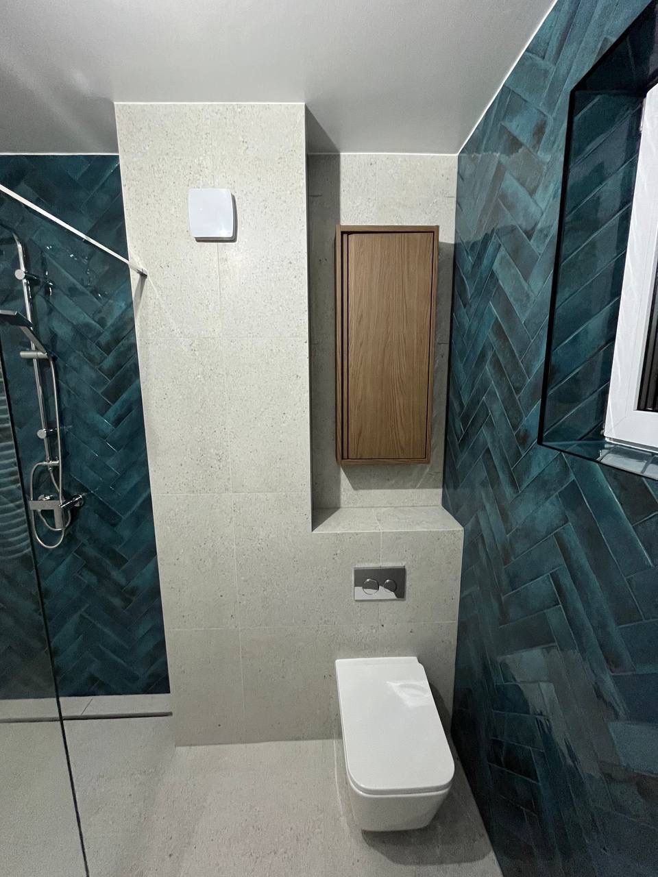 Nowoczesna łazienka z kabiną prysznicową, toaletą podwieszaną i szafką. Ściany wyłożone płytkami w odcieniach szarości i turkusu ułożonymi w jodełkę.