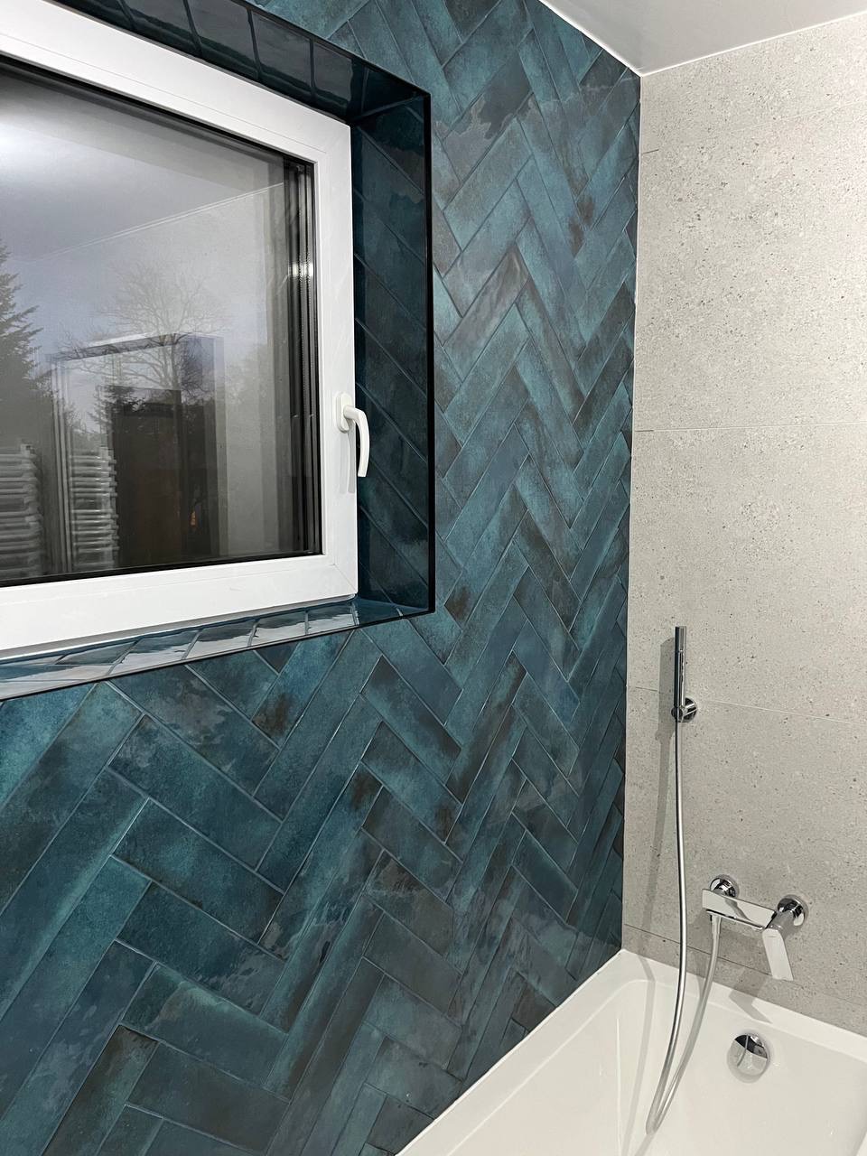 Wykończona łazienka: turkusowa ściana z płytek ułożonych w jodełkę, białe okno z widokiem na zewnątrz, szara ściana z baterią prysznicową nad wanną.