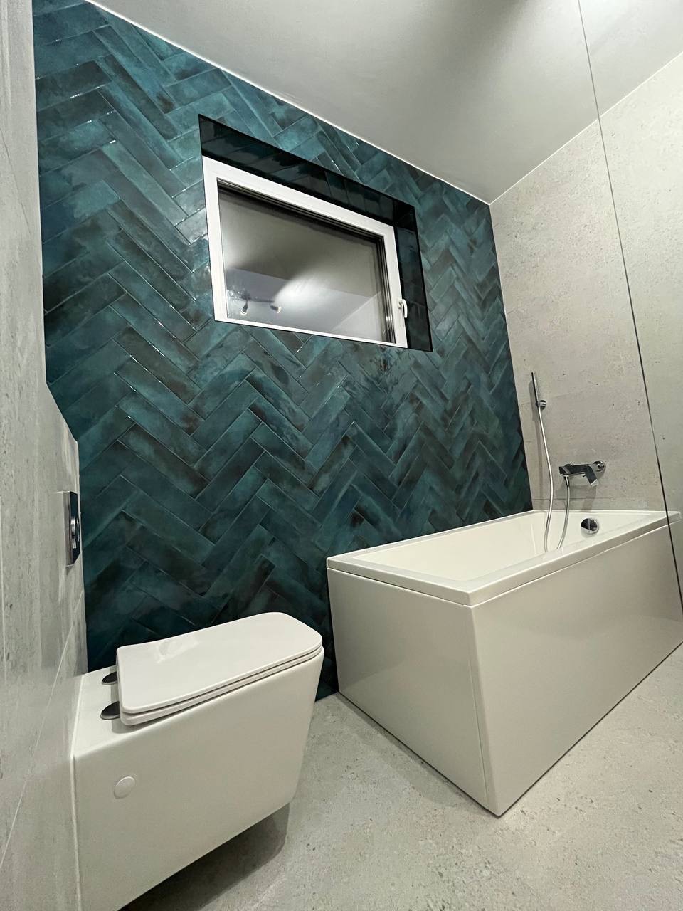 Nowoczesna łazienka z turkusową ścianą z płytek ułożonych w jodełkę, białą wanną i podwieszaną toaletą. Minimalistyczny styl, stonowana kolorystyka.