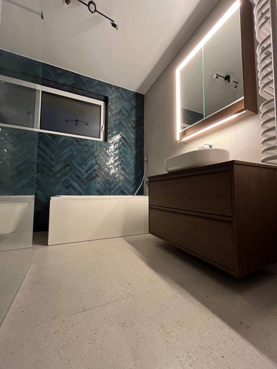 Nowoczesna łazienka z turkusową mozaiką ułożoną w jodełkę, białą wanną i szafką z umywalką podświetlaną lustrem. Minimalistyczny design.