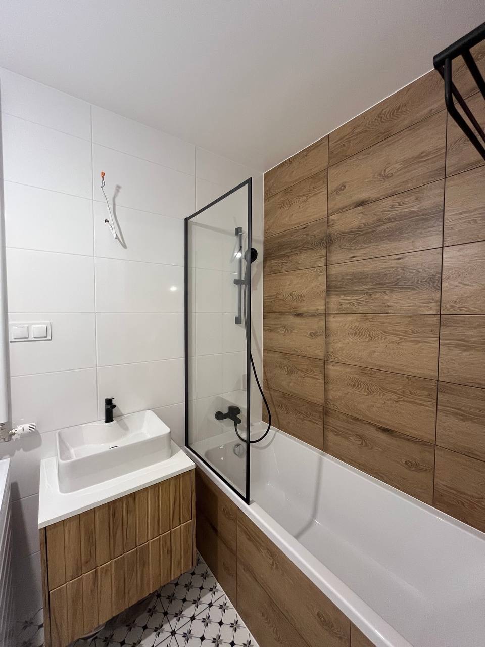 Nowoczesna łazienka z drewnopodobnymi płytkami, białą armaturą i czarnymi detalami, kabina prysznicowa z czarną ramą, wzorzysta podłoga.