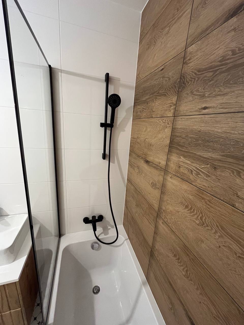 Nowoczesna łazienka: białe płytki, drewnopodobna okładzina, czarna armatura. Minimalistyczny design z akcentem na naturalne materiały i kontrastowe kolory.