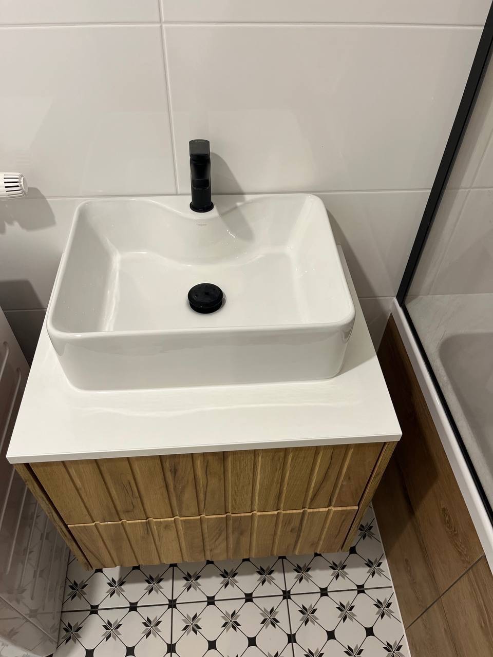 Nowoczesna umywalka nablatowa z czarną armaturą, osadzona na szafce z drewnianymi frontami. Białe płytki na ścianie, wzorzysta posadzka. Minimalistyczny design.