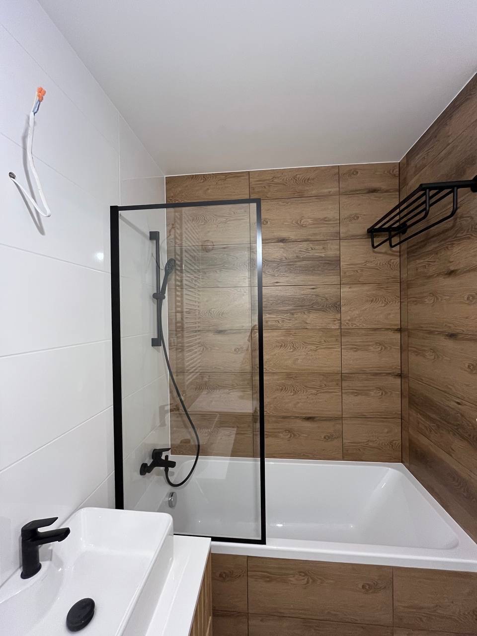 Nowoczesna łazienka z wanną za szklaną ścianką, białą umywalką i drewnopodobnymi płytkami. Widoczny czarny osprzęt i wystające przewody.