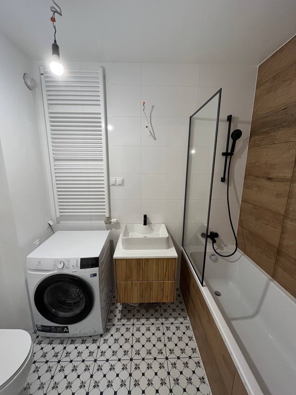 Wykończona łazienka z pralką, białą armaturą i wzorzystą podłogą. Kabina prysznicowa z czarną armaturą, umywalka z drewnopodobną szafką. Grzejnik drabinkowy.