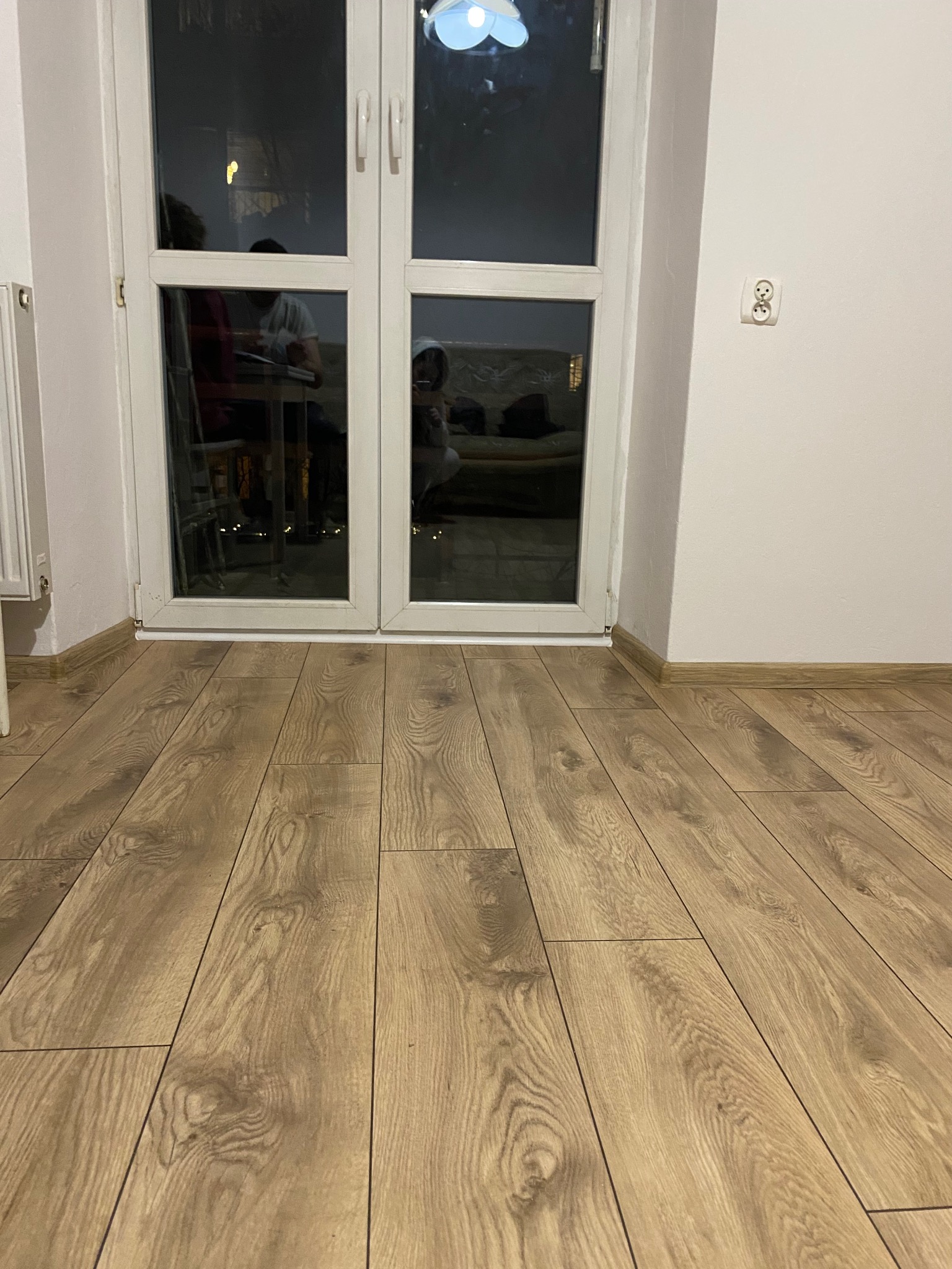 Jasne panele podłogowe ułożone wzdłuż białych drzwi balkonowych. Na ścianie widoczne gniazdko elektryczne. W tle odbicie osób w szybie.