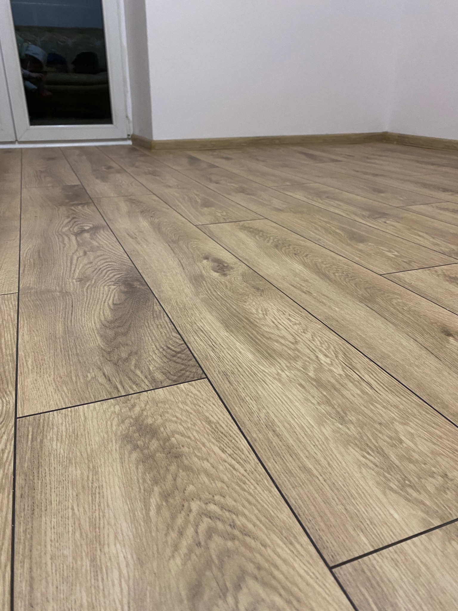 Jasne panele podłogowe ułożone w nowym pomieszczeniu, widoczne listwy przypodłogowe i okno z odbiciem. Ujęcie z niskiej perspektywy, akcentujące wzór drewna.