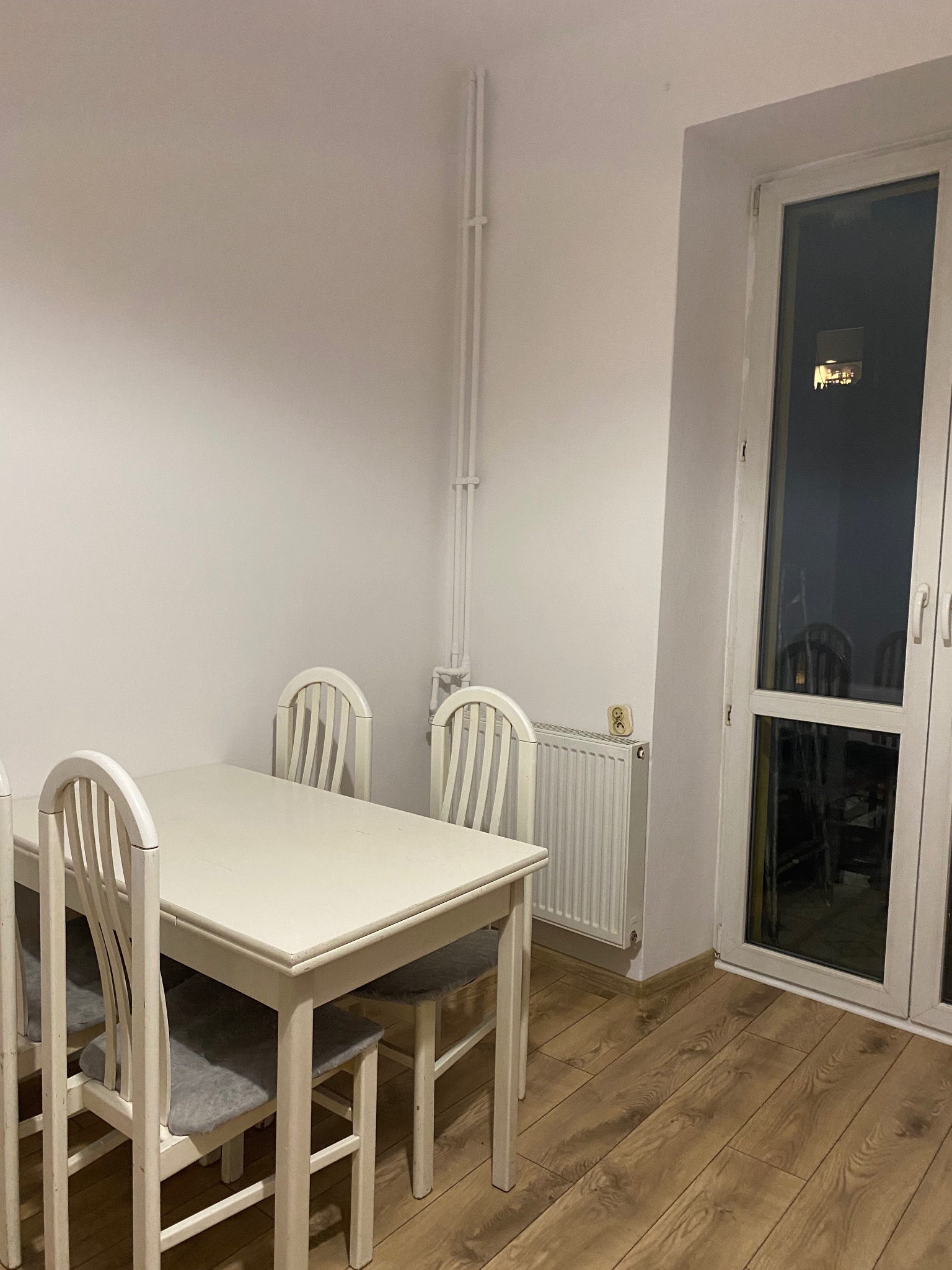 Jasne wnętrze jadalni po remoncie: biały stół z krzesłami, drewniana podłoga, grzejnik i okno balkonowe. Minimalistyczny design, świeżo odnowione ściany.