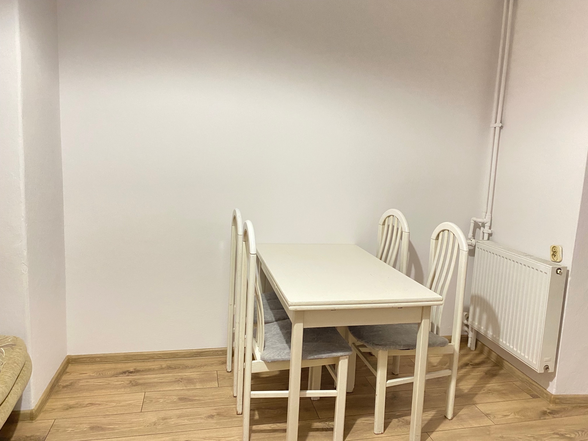 Jasne wnętrze z drewnianą podłogą, białymi ścianami i białym stołem z krzesłami. Widoczny grzejnik i rury przy ścianie. Minimalistyczny wystrój.