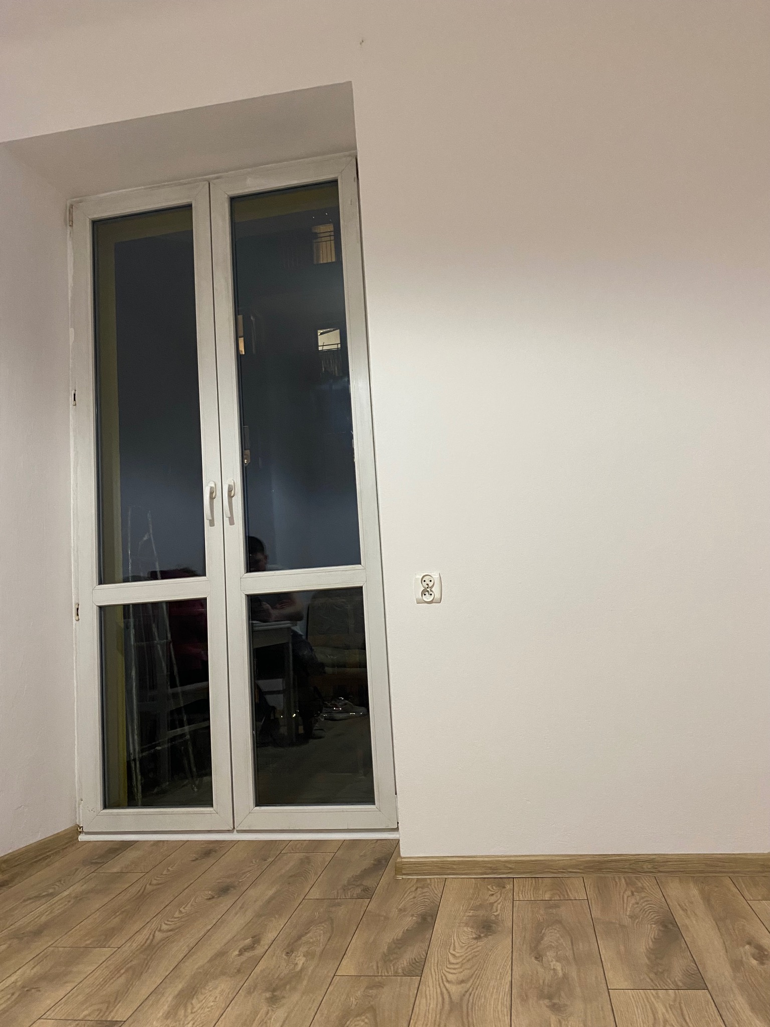 Jasne panele podłogowe w pokoju z białymi ścianami i oknem balkonowym. Widoczne gniazdko elektryczne. Minimalistyczny wystrój.