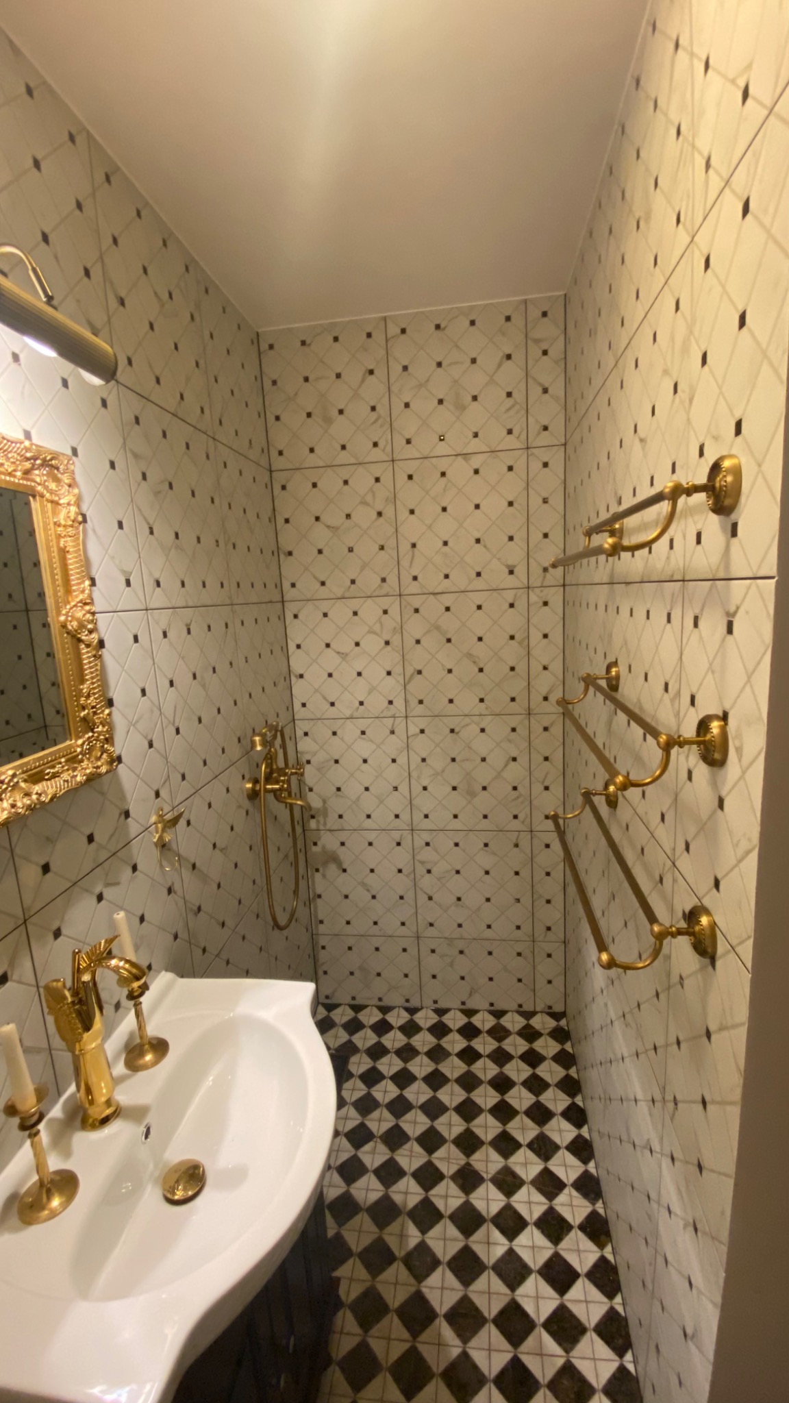 Elegancka łazienka w stylu vintage z białą umywalką, złotą armaturą i lustrem w ozdobnej ramie. Ściany i podłoga wyłożone płytkami w czarno-białą szachownicę.