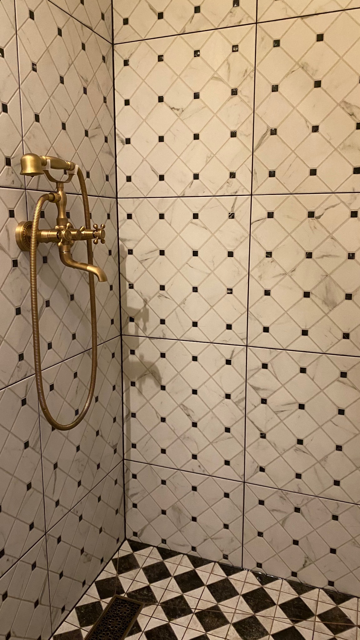 Wykończona kabina prysznicowa z biało-czarnymi płytkami w romby i złotą armaturą w stylu retro. Widoczny fragment podłogi z czarno-białą szachownicą.