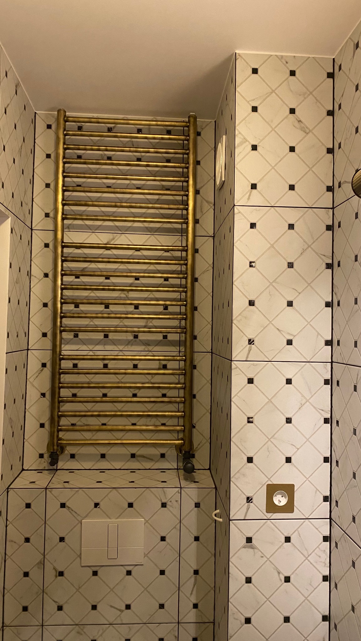 Wykończona łazienka z grzejnikiem drabinkowym w złotym kolorze na tle białych płytek z czarnymi akcentami. Widoczny panel sterowania spłuczką WC.