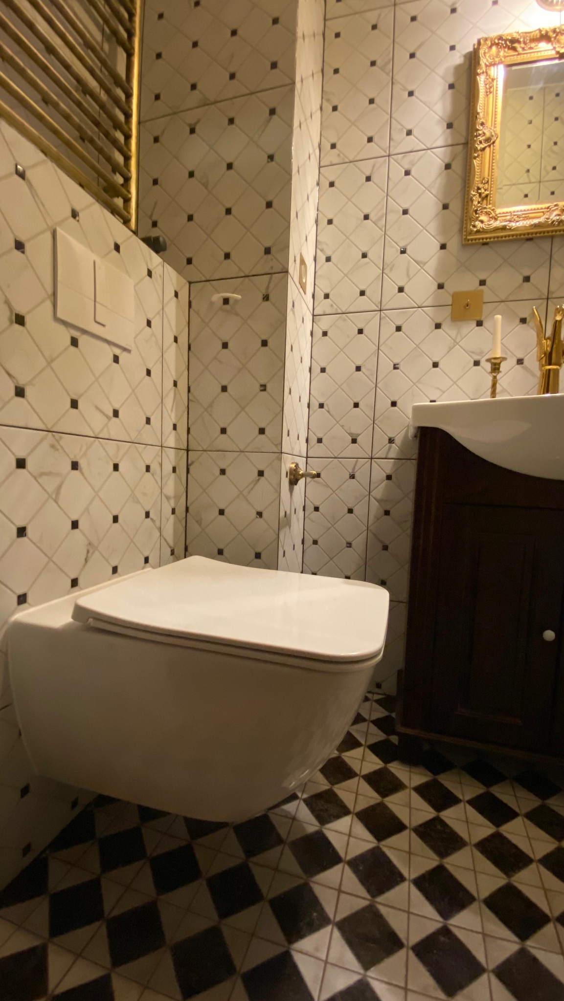 Elegancka łazienka z białą toaletą, umywalką w ciemnym drewnie i czarno-białą podłogą w kratkę. Złote akcenty na grzejniku i lustrze dodają luksusowego charakteru.