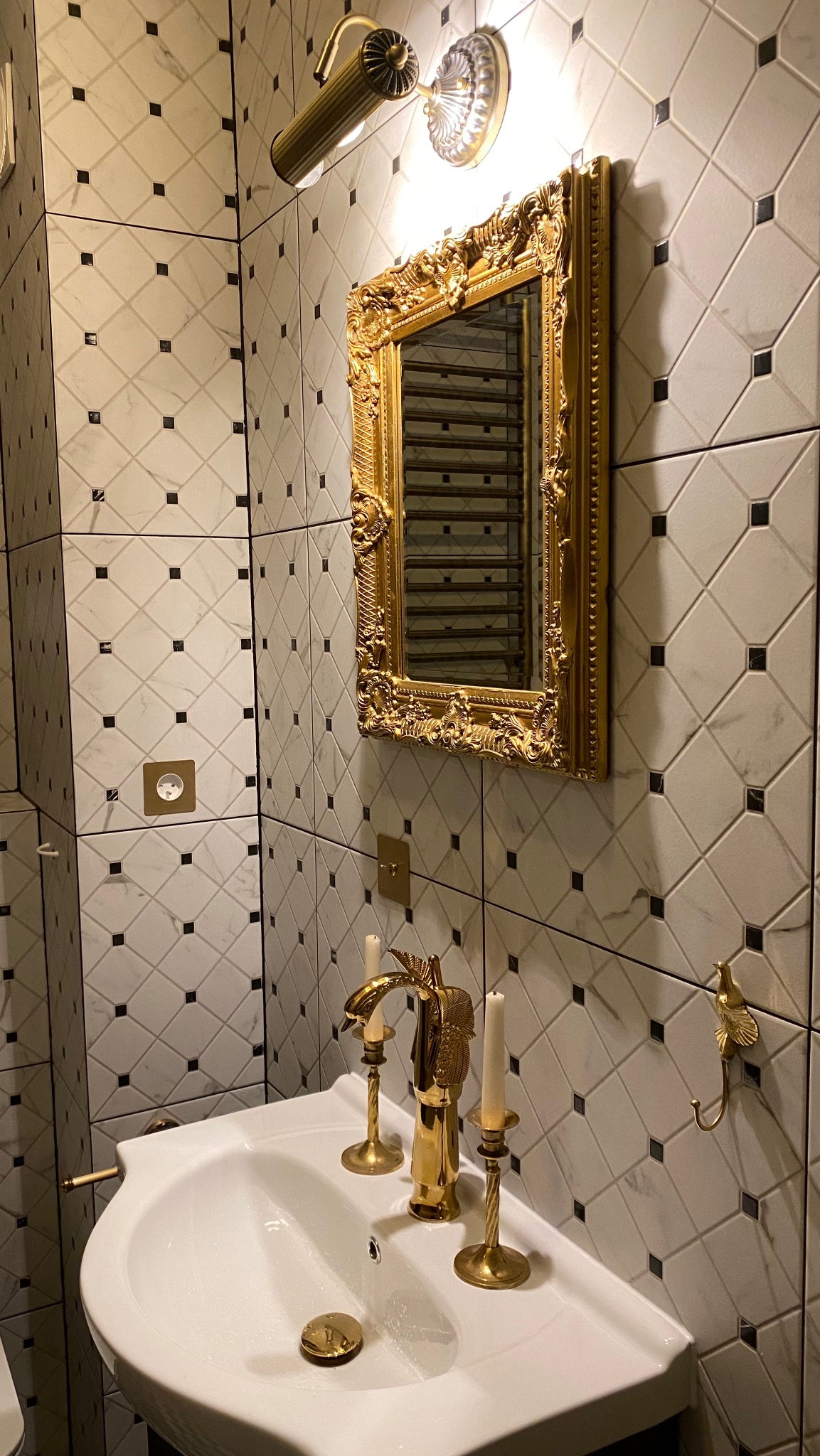 Elegancka łazienka: biała umywalka z złotą armaturą i świecznikami, lustro w bogato zdobionej, złotej ramie, płytki w biało-czarną szachownicę i złote dodatki.