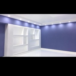 Home & Trade Tomasz Gliński - Wnętrze pokoju z wbudowaną, białą szafką z półkami na ścianie pomalowanej na fioletowo, oświetlone punktowymi lampami sufitowymi.