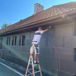 Home & Trade Tomasz Gliński - Pracownik na drabinie montuje grafitowe płyty izolacyjne na elewacji domu jednorodzinnego w słoneczny dzień, widoczne okna i dachówka.