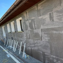 Home & Trade Tomasz Gliński - Elewacja budynku w trakcie ocieplania, widoczne płyty izolacyjne z szarego styropianu, profile startowe, okna przygotowane do montażu parapetów i rusztowanie na ziemi.