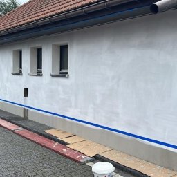 Home & Trade Tomasz Gliński - Ściana zewnętrzna domu przygotowywana do malowania: świeżo nałożona szpachla, zabezpieczona taśmą malarską przy oknach i cokole, drabina i wiadro z farbą w pobliżu.