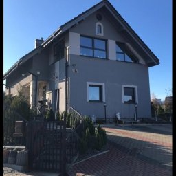 Home & Trade Tomasz Gliński - Dwukondygnacyjny dom jednorodzinny z szarą elewacją, czarną stolarką okienną i brukowanym podjazdem w słoneczny dzień.