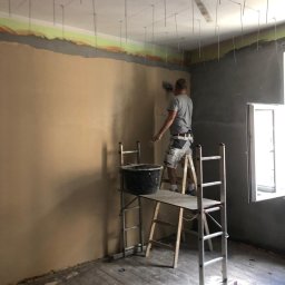Home & Trade Tomasz Gliński - Mężczyzna na rusztowaniu nakłada gładź na ścianę w pomieszczeniu z przygotowanym sufitem pod zabudowę z płyt karton-gips, widoczny kubeł z materiałem i drewniana podłoga.