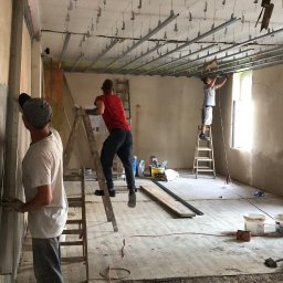 Home & Trade Tomasz Gliński - Montaż metalowej konstrukcji sufitu podwieszanego z widocznymi elementami mocującymi w pomieszczeniu w trakcie remontu w Piekarach Śląskich. Trzech mężczyzn pracuje na drabinach, mocując profile...