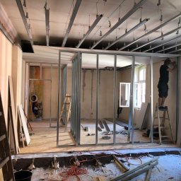 Home & Trade Tomasz Gliński - Montaż metalowej konstrukcji ścian działowych z profili w trakcie remontu mieszkania, widoczne profile sufitowe, płyty OSB, drabina i pracownik na drabinie przy oknie.