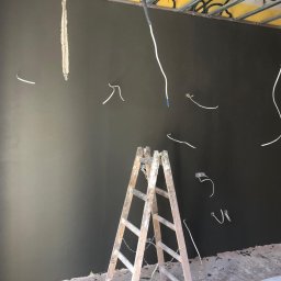 Home & Trade Tomasz Gliński - Świeżo nałożona, ciemnoszara gładź na ścianie z wystającymi przewodami elektrycznymi, drewniana drabina rozstawiona na środku pomieszczenia, widoczne elementy konstrukcji sufitu.