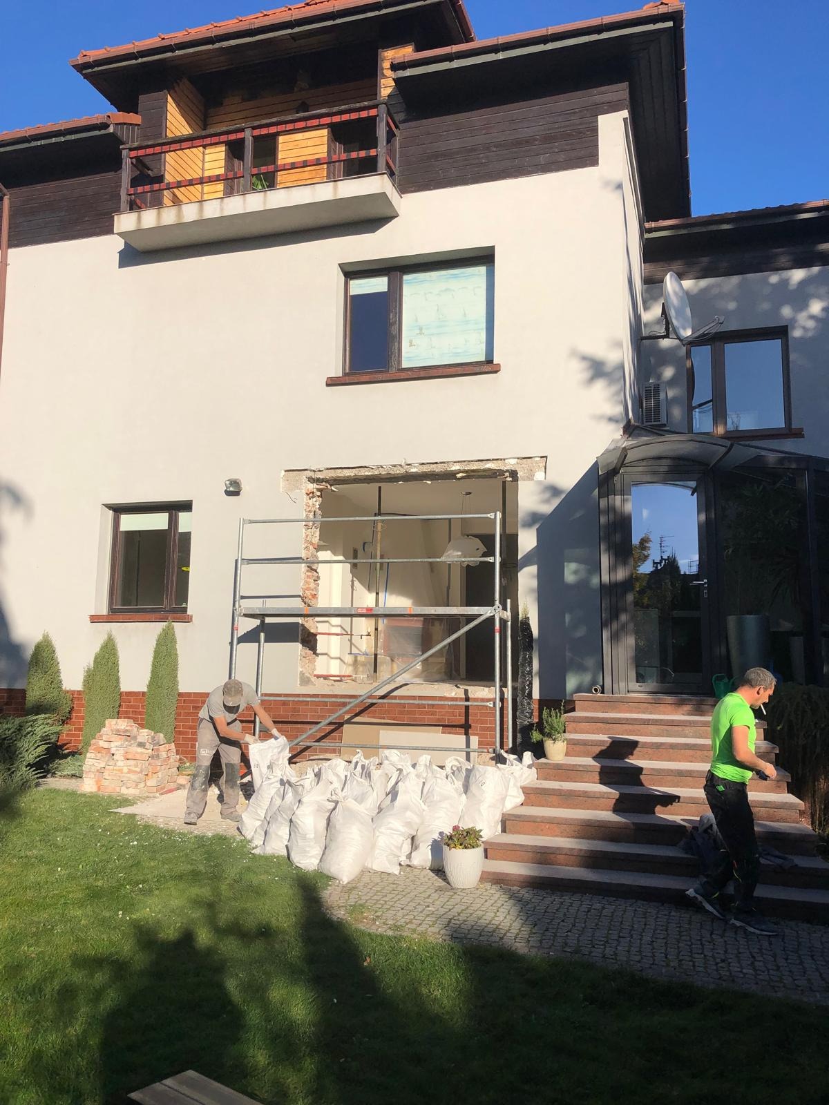 Pracownik w trakcie prac murarskich przy wykuwaniu otworu okiennego w elewacji domu, z przygotowanymi workami z gruzem i cegłami, widoczne rusztowanie.