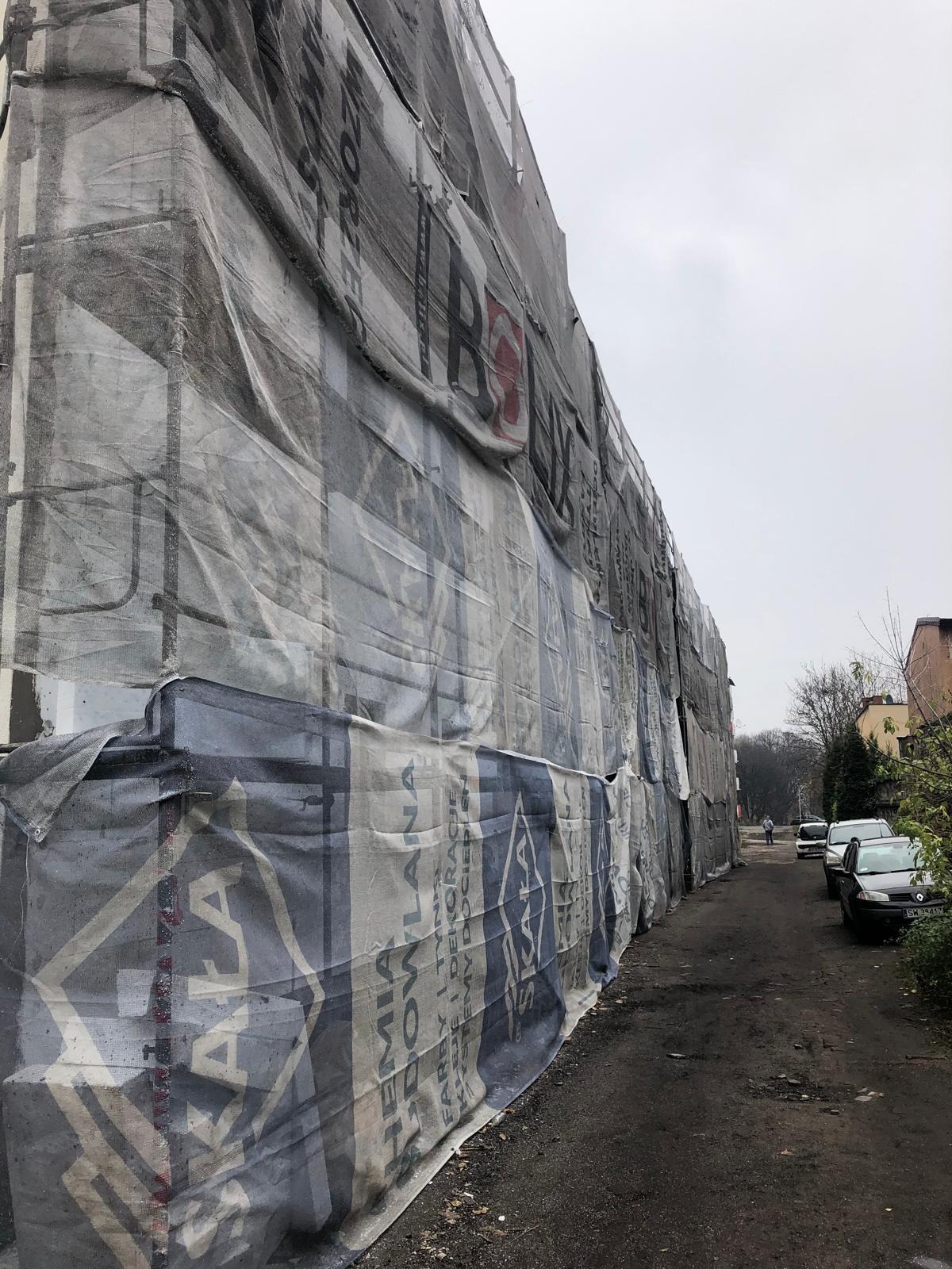 Budynek w trakcie remontu, osłonięty siatką z reklamą materiałów budowlanych, widok z boku ulicy.