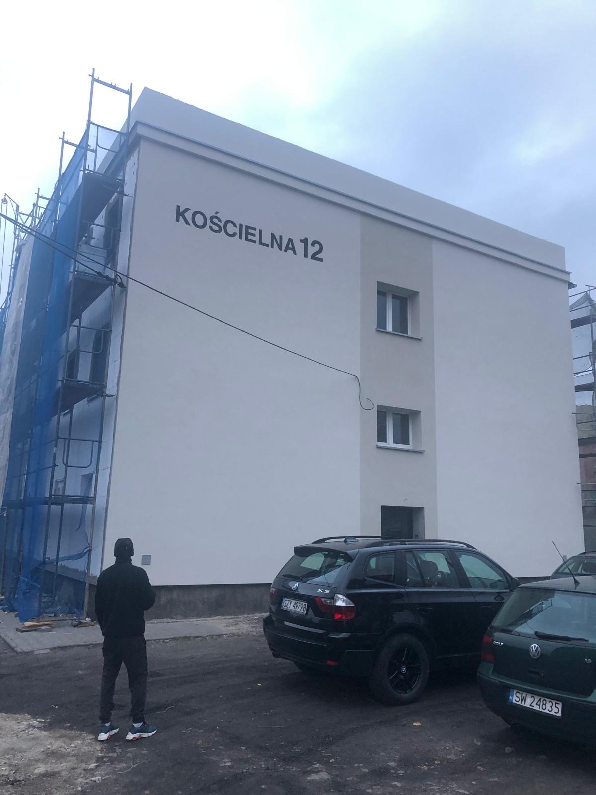 Budynek z nową, jasną elewacją z napisem 'Kościelna 12', widoczny fragment rusztowania z siatką ochronną, zaparkowane samochody i postać mężczyzny obserwującego budynek.
