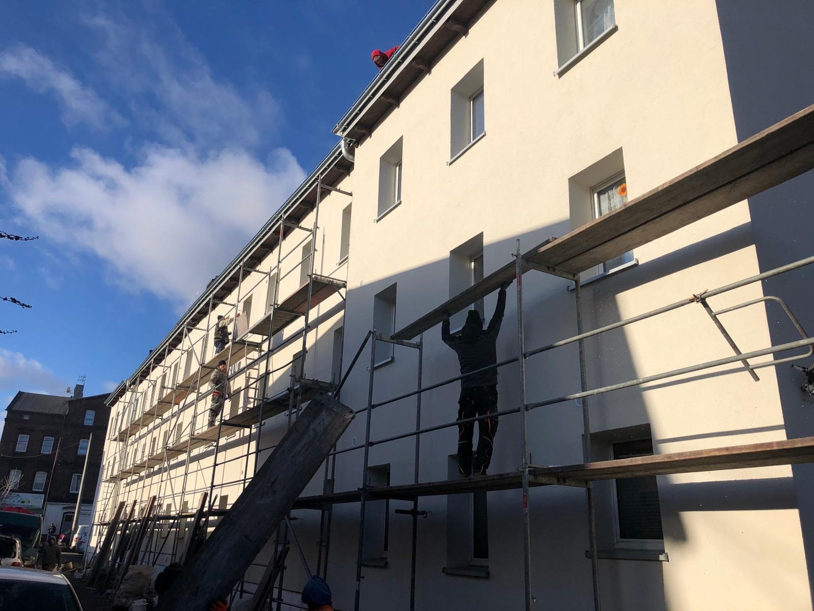 Pracownicy na rusztowaniu podczas odnawiania elewacji budynku wielorodzinnego, widoczne świeżo pomalowane ściany w jasnym kolorze, błękitne niebo z chmurami w tle.