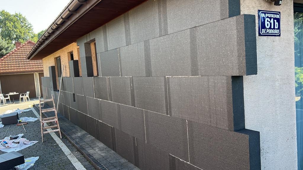 Ocieplanie ściany domu styropianem grafitowym, widoczna drabina oparta o budynek i tabliczka z adresem.