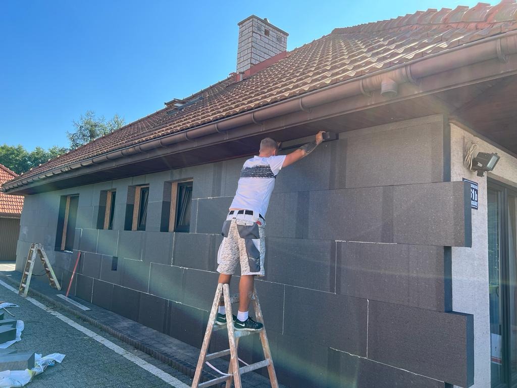 Pracownik na drabinie montuje grafitowe płyty izolacyjne na elewacji domu jednorodzinnego w słoneczny dzień, widoczne okna i dachówka.