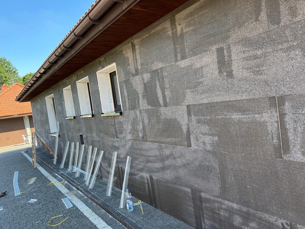 Elewacja budynku w trakcie ocieplania, widoczne płyty izolacyjne z szarego styropianu, profile startowe, okna przygotowane do montażu parapetów i rusztowanie na ziemi.