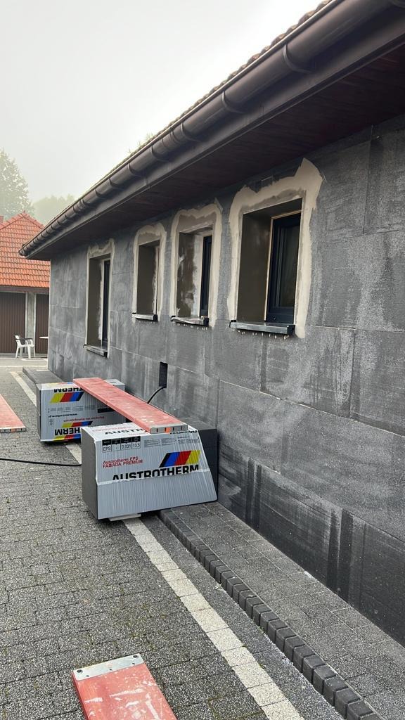 Ocieplenie budynku styropianem Austrotherm, widoczne puszki okienne przygotowane do osadzenia okien, elewacja pokryta warstwą izolacyjną, fragment dachu z rynną.