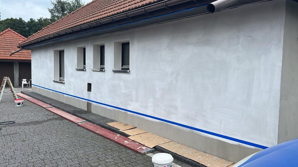 Ściana zewnętrzna domu przygotowywana do malowania: świeżo nałożona szpachla, zabezpieczona taśmą malarską przy oknach i cokole, drabina i wiadro z farbą w pobliżu.