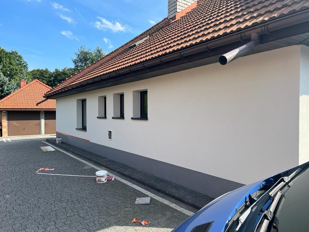 Świeżo pomalowana ściana domu jednorodzinnego z widocznymi trzema oknami, z zaznaczonym odcięciem kolorów i wiadrem farby na brukowanym podjeździe.