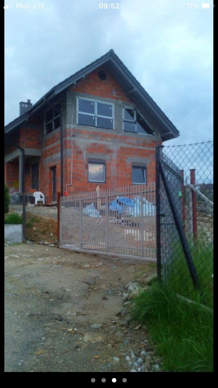 Niedokończony dom jednorodzinny z czerwonej cegły, z zamontowanymi oknami, widoczny od frontu przez metalową bramę i siatkę ogrodzeniową.