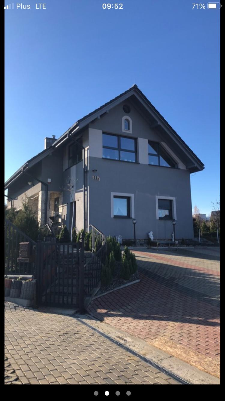 Dwukondygnacyjny dom jednorodzinny z szarą elewacją, czarną stolarką okienną i brukowanym podjazdem w słoneczny dzień.