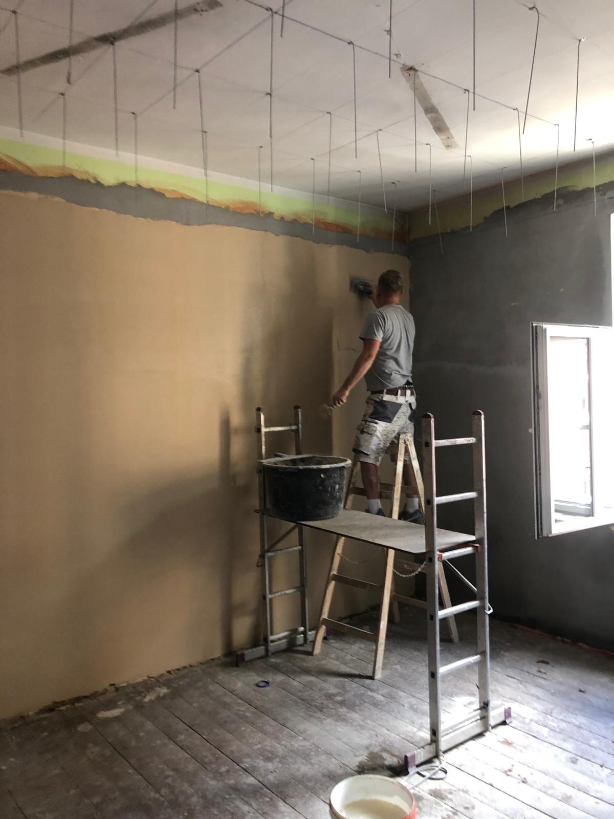 Mężczyzna na rusztowaniu nakłada gładź na ścianę w pomieszczeniu z przygotowanym sufitem pod zabudowę z płyt karton-gips, widoczny kubeł z materiałem i drewniana podłoga.