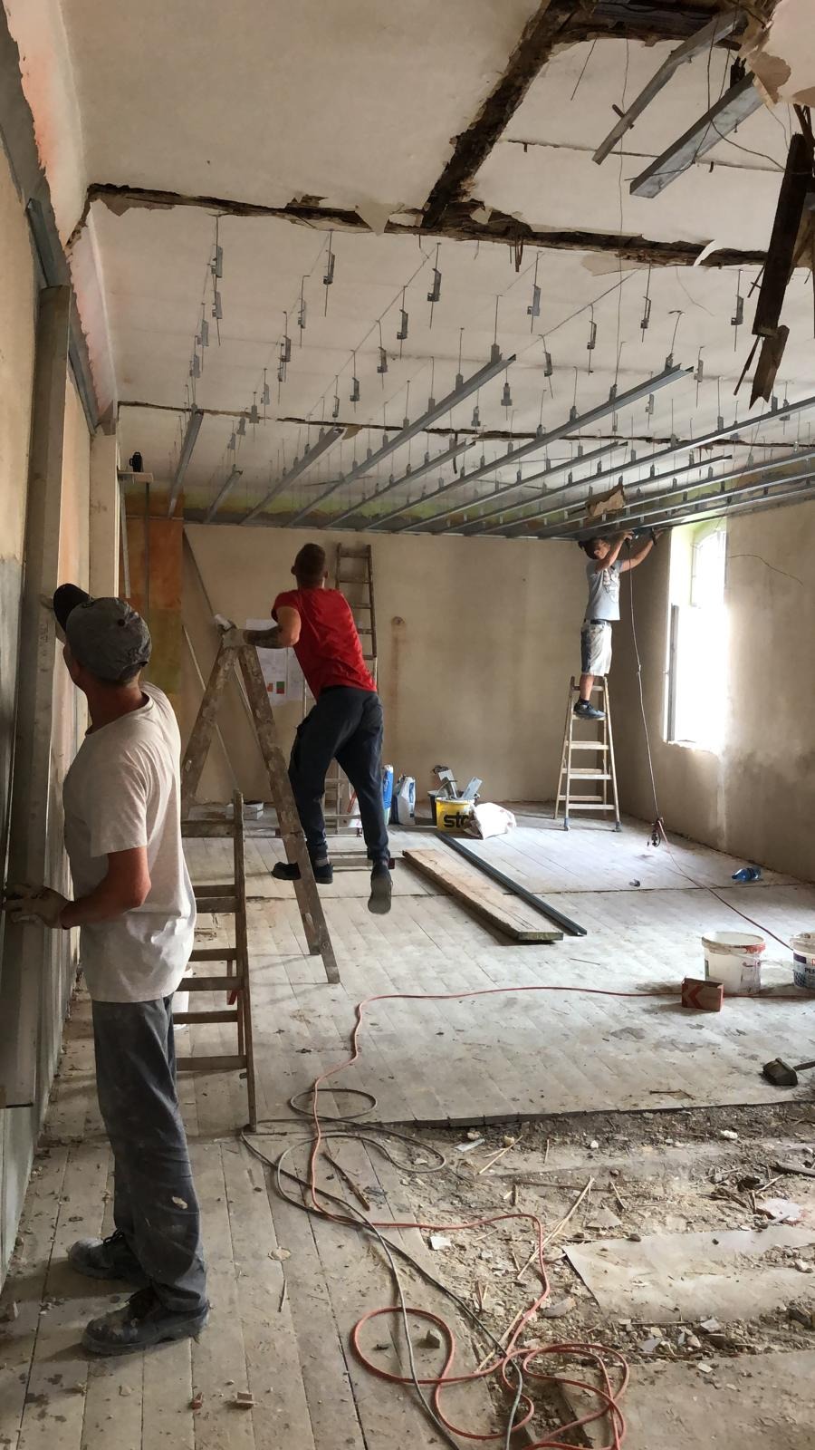 Montaż metalowej konstrukcji sufitu podwieszanego z widocznymi elementami mocującymi w pomieszczeniu w trakcie remontu w Piekarach Śląskich. Trzech mężczyzn pracuje na drabinach, mocując profile...