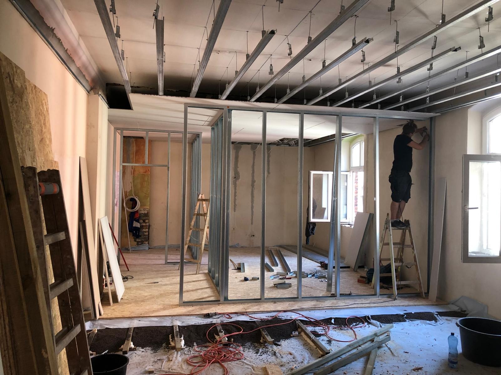 Montaż metalowej konstrukcji ścian działowych z profili w trakcie remontu mieszkania, widoczne profile sufitowe, płyty OSB, drabina i pracownik na drabinie przy oknie.