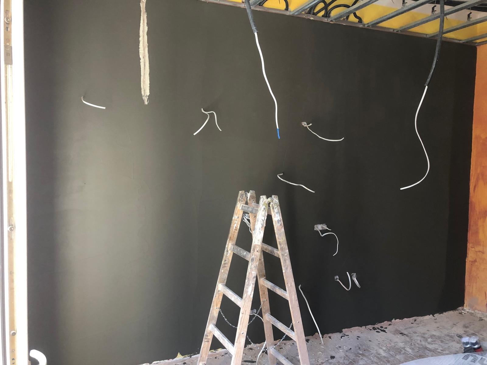 Świeżo nałożona, ciemnoszara gładź na ścianie z wystającymi przewodami elektrycznymi, drewniana drabina rozstawiona na środku pomieszczenia, widoczne elementy konstrukcji sufitu.